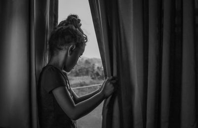 Side view of girl looking through window while standing at home