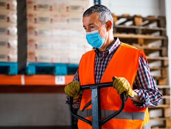 Focused adult male worker wearing high visibility vest and face mask holding trolley cart handle and looking down while working in contemporary storehouse