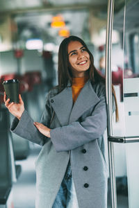 A happy brunette with coffee in her hands stands on the train, looks into the distance and smiles