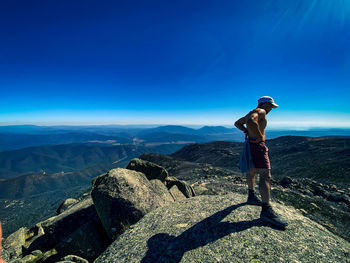 Man standing on rock looking at mountains against blue sky