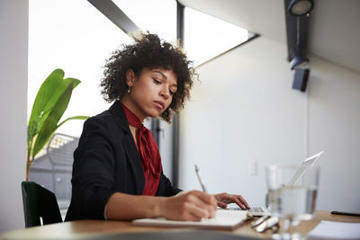 Confident young female financial advisor writing on diary while sitting with laptop at desk in office