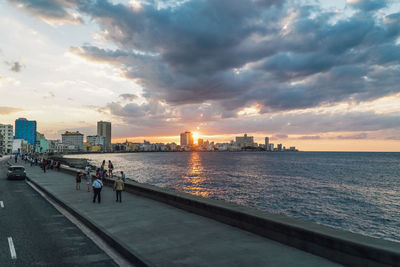 People on city by buildings against sky during sunset