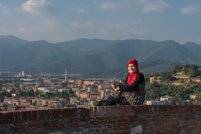 Woman sitting on mountain by cityscape against sky