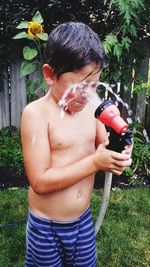 Shirtless boy splashing water with hose while standing in backyard