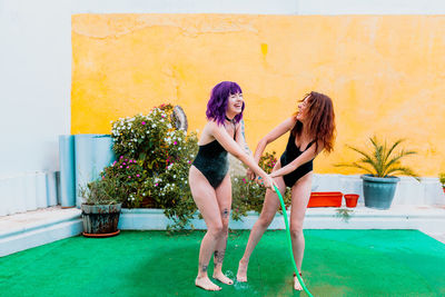 Two happy laughing women in swimsuit playing under water droplets from a hose at backyard