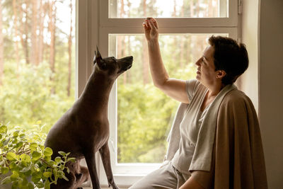 Middle age beautiful woman sitting on windowsill with her dog. 50-year-old woman