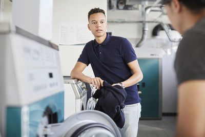 Smiling man listening to male friend while doing laundry at laundromat