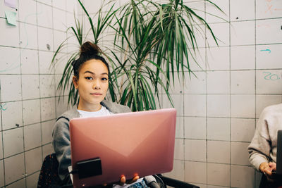 Portrait of confident young female hacker using laptop while sitting against tile wall at small creative office