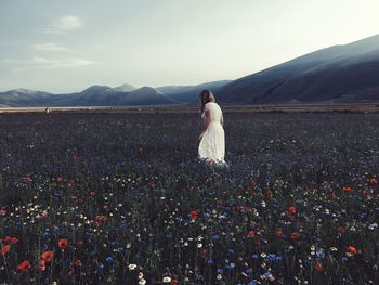 Woman walking amidst flowering plants on field by mountains against sky