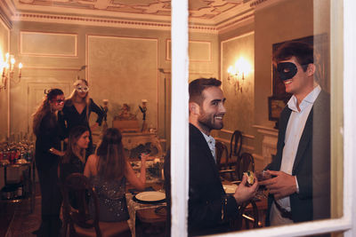 Through window of group of friends in elegant wear and masquerade masks chatting while having dinner together in restaurant