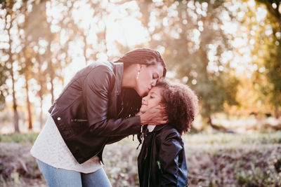 Mother kissing daughter while standing against trees