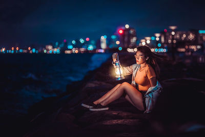 Woman sitting on thr sea wall holding a vintage lamp with city lights on the background.