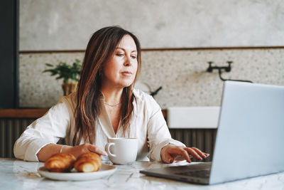 Adult smiling brunette woman having breakfast with opened laptop in kitchen at home