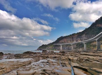 Sea cliff bridge by rocky mountains against cloudy sky