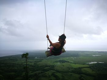 Woman wearing hat looking over shoulder while swinging on swing against cloudy sky