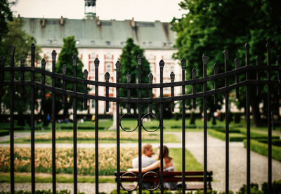 Rear view of couple sitting on bench at park seen through fence