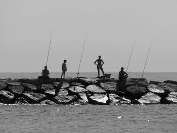 People with fishing rods standing on rocks amidst sea against clear sky during sunny day