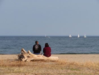 Rear view of couple sitting on log at beach against sky