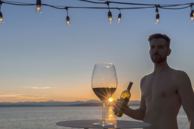 Wineglass on table with man holding bottle while standing at beach against sky during sunset