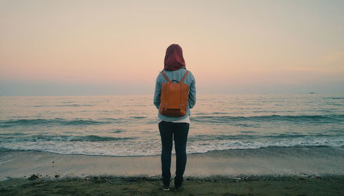 Rear view of woman standing at sea shore against sky during sunset