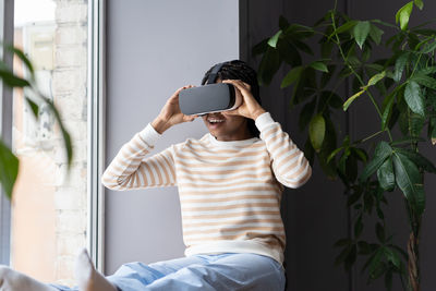 Young amazed excited afro girl wearing vr helmet relaxing with virtual reality on windowsill at home