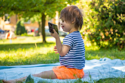 Side view of cute boy holding smart phone sitting at park