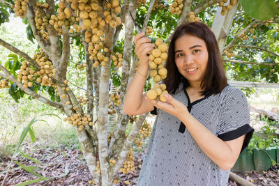 Portrait of smiling young woman showing fruits against tree