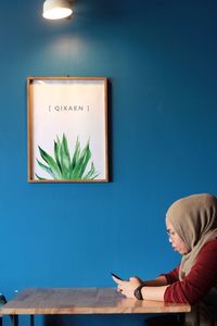 Rear view of woman sitting by potted plant on table against wall