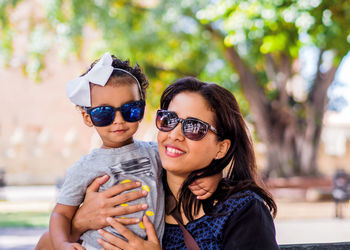 Portrait of smiling mother and daughter wearing sunglasses against trees at park