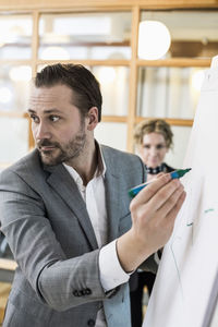 Mid adult businessman writing on flip chart with female colleague in background at office