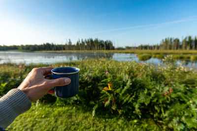 Close-up of hand holding drink by lake against sky