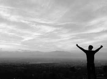 Rear view of man with arms raised looking at cityscape against sky