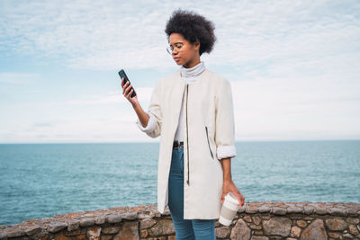 Woman using mobile phone while standing against sea