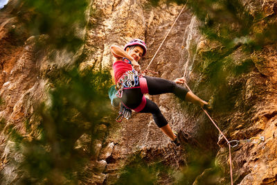 Low angle full length of active senior woman climbing on rocky cliff against sky