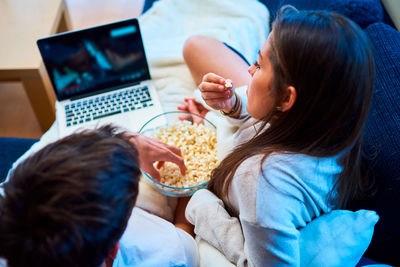 Cheerful young man and woman in casual wear eating popcorn and watching film on laptop while resting together on cozy sofa at home