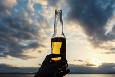 Cropped hand holding beer bottle against sky during sunset