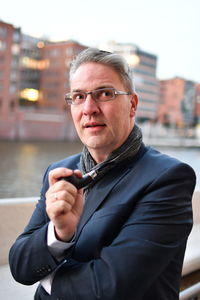 Businessman looking away while holding smoking pipe while standing against buildings and sky