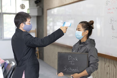 Man wearing flu mask checking temperature of woman holding slate in classroom