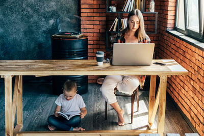 A working mother on a laptop at a table, a child sitting on the floor reading a book.