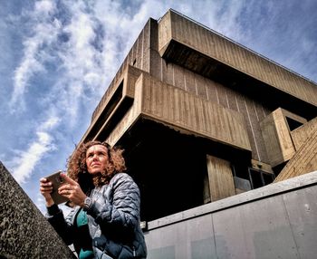 Low angle view of woman using mobile phone while standing by retaining wall against cloudy sky