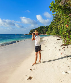 Young man in white t-shirt standing on sandy beach, taking photos.