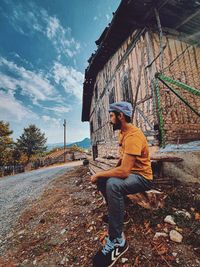 Side view of young man sitting by hut against sky