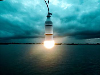 Illuminated light bulb hanging against sea at dusk