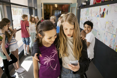 Girls using mobile phone with friends in corridor at school