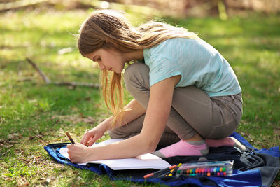 Young adolescent girl writes or colors in a notebook or journal on a blanket on the grass sunny day