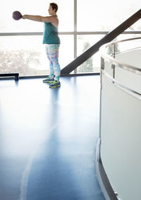 Side view of woman lifting kettlebell while standing by window in gym