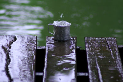 Water falling in metallic mug on wooden plank by lake