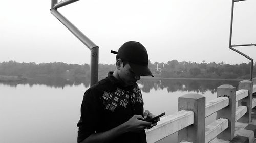 Side view of young man using phone standing on bridge against lake