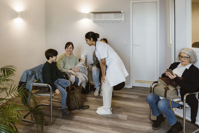 Female doctor with hands on knee talking to boy sitting with mother in waiting room