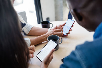 Group of cropped unrecognizable multiracial coworkers surfing cellphones while sitting at table with hot drinks during break in modern light cafe